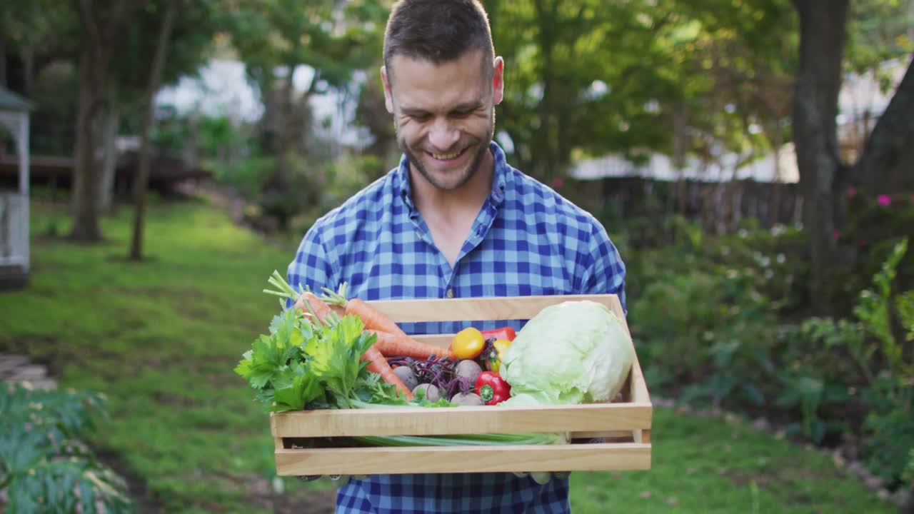 hombre caucásico sonriente de pie en el jardín sosteniendo caja de verduras