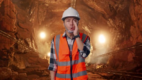 asian male engineer with safety helmet yelling with hand over mouth while standing in underground mine tunnel