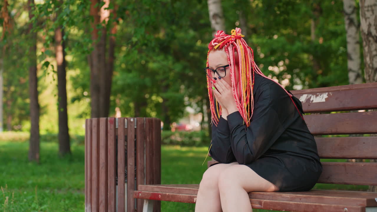 Mujer blanca sentada en un banco con rastas llamativas, con las manos en la cara en una pose de frustración, ambiente tranquilo de parque frondoso, papelera cerca, conjunto negro de moda, momento emocional de bloqueo creativo