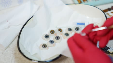 few eye prosthesis in medical bowl. medical specialist in red gloves painting artificial eye in the laboratory. close-up.