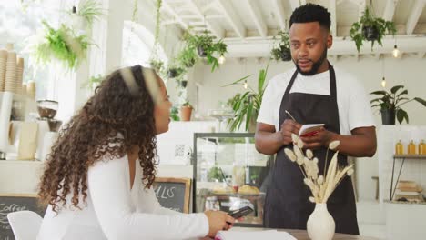 Glücklicher-Afroamerikanischer-Männlicher-Barista,-Der-Im-Café-Die-Bestellung-Einer-Biracial-Weiblichen-Kundin-Entgegennimmt