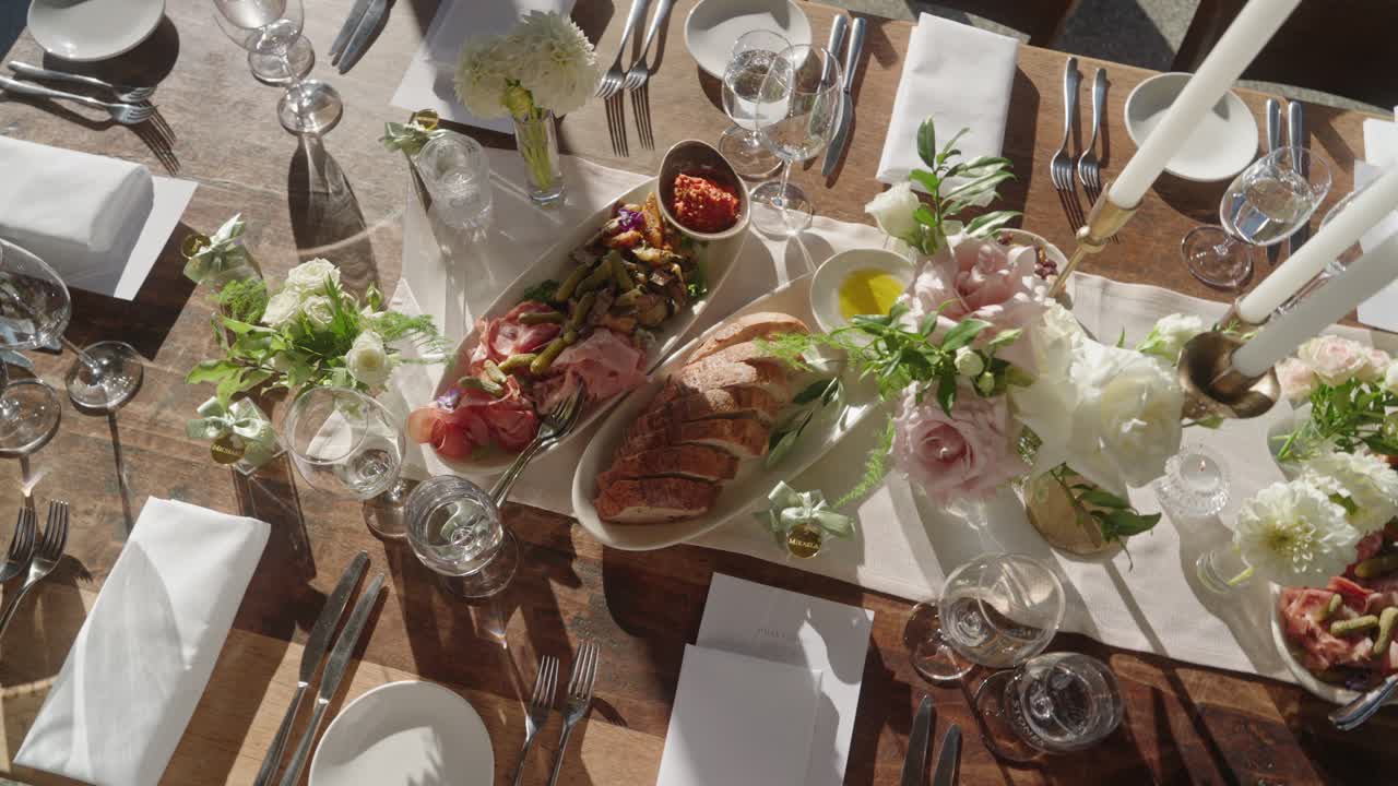 Top down view of an elegant wedding dining table with food, sliced bread, water in glasses, cutlery, napkins, floral centerpiece, candles, and plates arranged neatly
