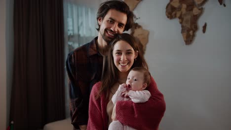 Portrait-of-a-happy-brunette-man-with-stubble-along-with-his-brunette-wife-and-little-baby-daughter-in-a-pink-overalls-posing-in-a-modern-apartment