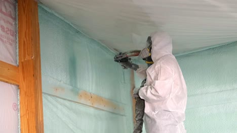 trabajador con respirador y traje tyvek rocía aislamiento de espuma de celda cerrada cerca del techo de una pared exterior recién construida