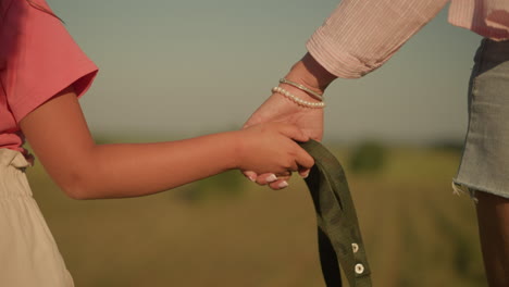 close-up of child and adult holding hands while gripping rope together, adult wears pearl bracelet, adding touch of elegance against background of vast, open farmland under warm sunlight