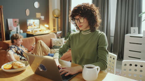 asian woman working on laptop while partner relaxing on sofa at home