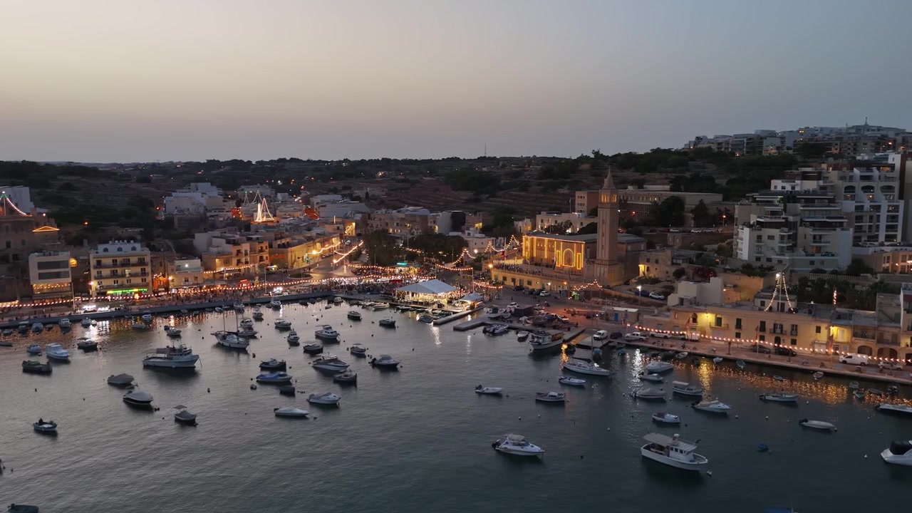 Marsaskala-bukten, Maltas församlingskyrka och marina med förtöjda båtar, lägenheter vid vattnet och upplysta byfestgator i skymningen, fångat med drönare