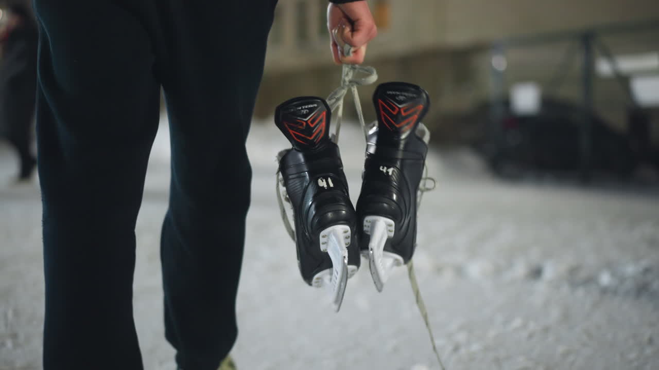 Back view of skater walking on snowy ground holding black ice skating boots by lace toward building in cold outdoor setting, with soft focus on icy surface and winter atmosphere