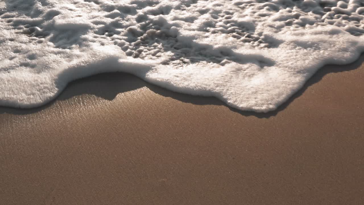 Close up footage capturing gentle sea waves reaching the coast and forming white foamy patterns on the sandy beach.