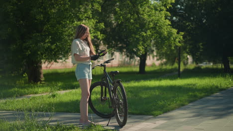 woman in gloves removes green air pump from bicycle tire, contemplating her work, surrounding area includes lush green trees and distant building