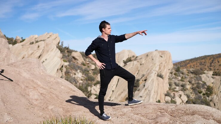 A young attractive fit man at the top of a mountain peak hike looks over the cliff at the landscape and points in slow motion