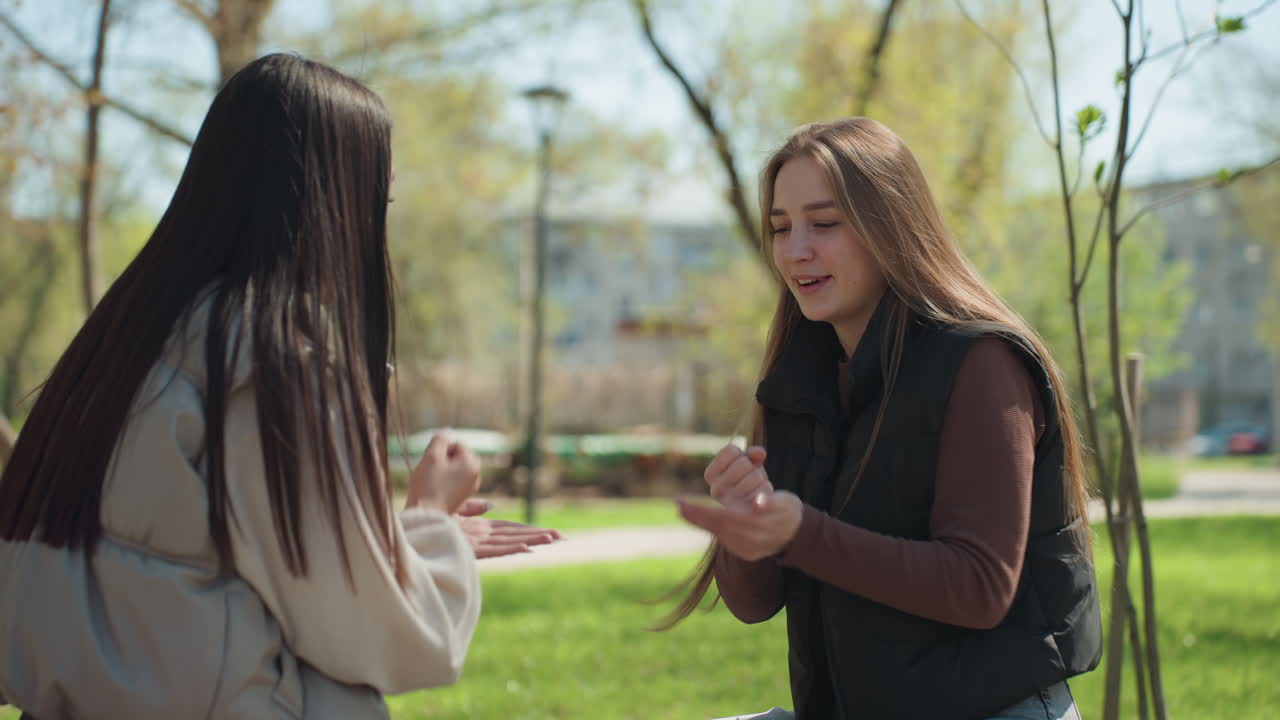 banco del parque, amigos, personas de ascendencia asiática y caucásica jugando a piedra, papel o tijera, tarde soleada de primavera con risas y gestos divertidos, dos mujeres jóvenes con chaquetas informales decidiendo el resultado, caras cercanas, vegetación y abedules