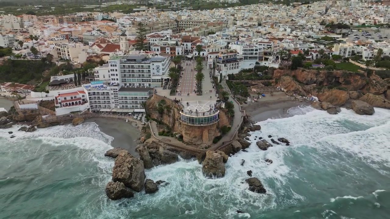 Luftaufnahme einer Drohne mit Rückwärtsbewegung und nach oben geneigter Kamera, die das Stadtzentrum von Nerja, den Aussichtspunkt Balcón de Europa, die palmenbestandene Promenade und grüne Berge im Hintergrund bei bewölktem Tageslicht zeigt.