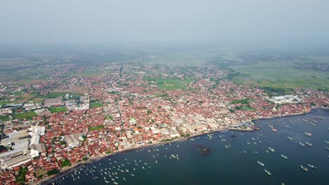 the aerial footage displays muncar harbor filled with traditional boats, nestled alongside densely populated residential areas and fish industrial zones