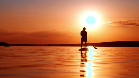silhouette of a man in cap standing on the sup board in the river. setting sun colors everything orange. low angle view.
