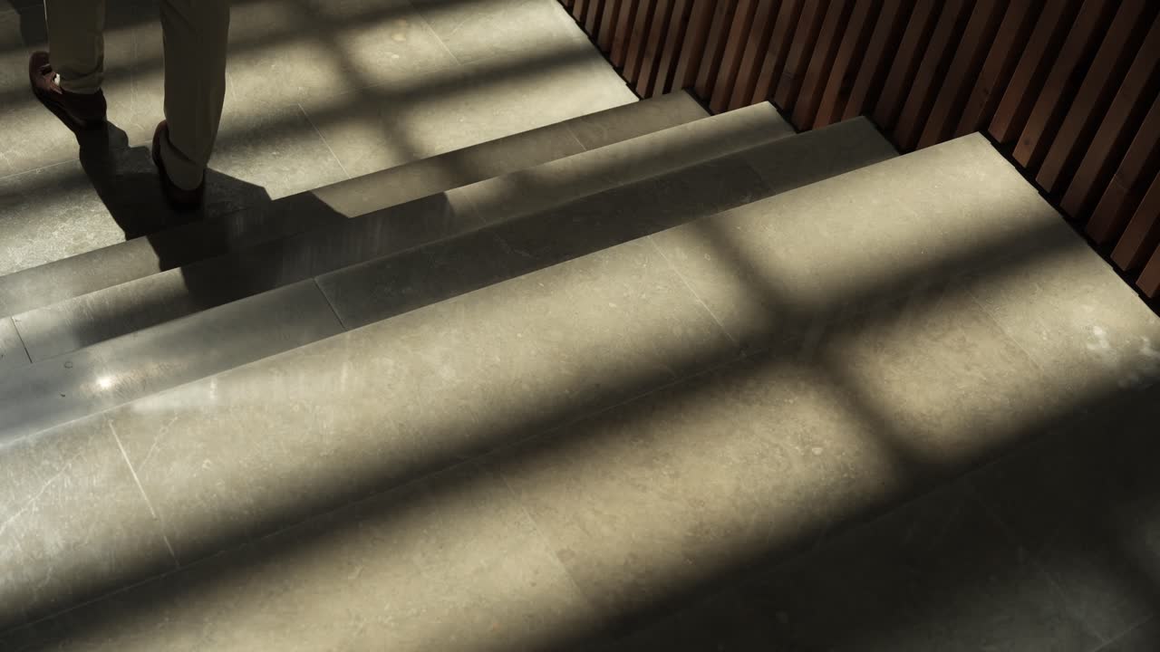 Cena em câmera lenta de um homem descendo escadas iluminadas pelo sol, projetando uma sombra alongada nos degraus.