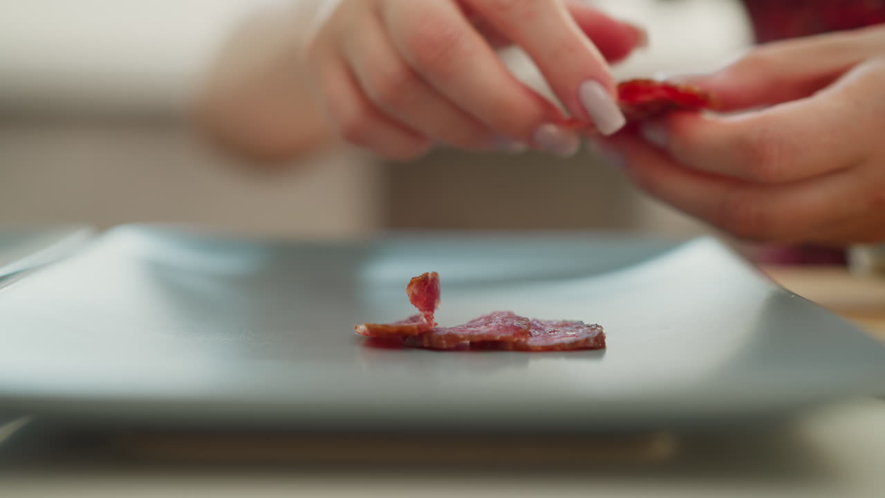 Pastry chef hands arranging single thin sausage slice on dark tray beside chopped meat pile on wooden board and whole sausage link on white table, soft natural light highlights texture and color
