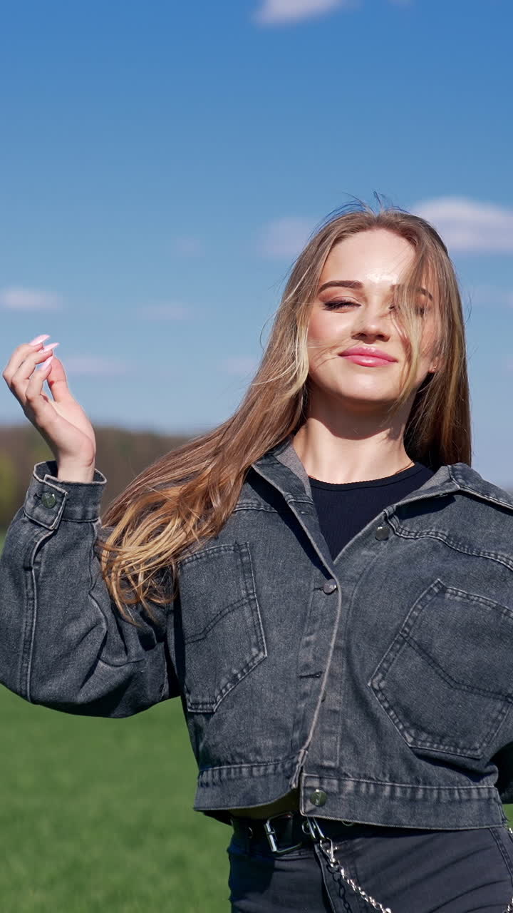 Happy girl on a meadow. Attractive young model with lovely face in denim suit posing on camera and smiles. Portrait of a young woman in nature in a sunny day. Vertical video