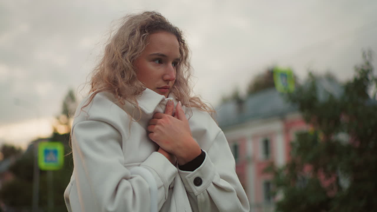 Shivering lady pulling white coat tightly around herself trying to stay warm in cold weather, with blurred background featuring colorful building, green trees, signpost, and cloudy sky
