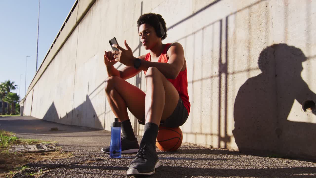 hombre afroamericano en forma en la ciudad, usando un teléfono inteligente con auriculares inalámbricos, sentado en el baloncesto