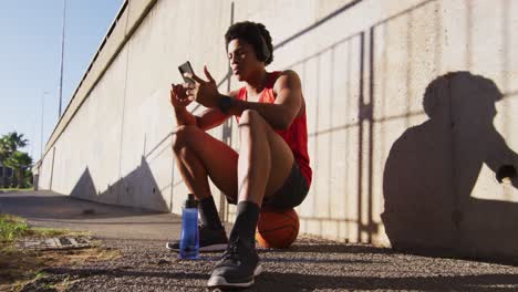 fit african american man in city, using smartphone with wireless headphones, sitting on basketball