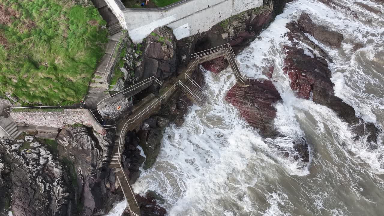 Drone video looking straight down at the waves crashing against the walk walk and rocks beside Youghal Lighthouse in Youghal, East Cork, Ireland