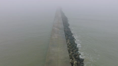 aerial establishing view of port of liepaja concrete pier, baltic sea coastline , foggy day with dense mist, moody feeling, big storm waves splashing, wide birdseye drone shot moving forward