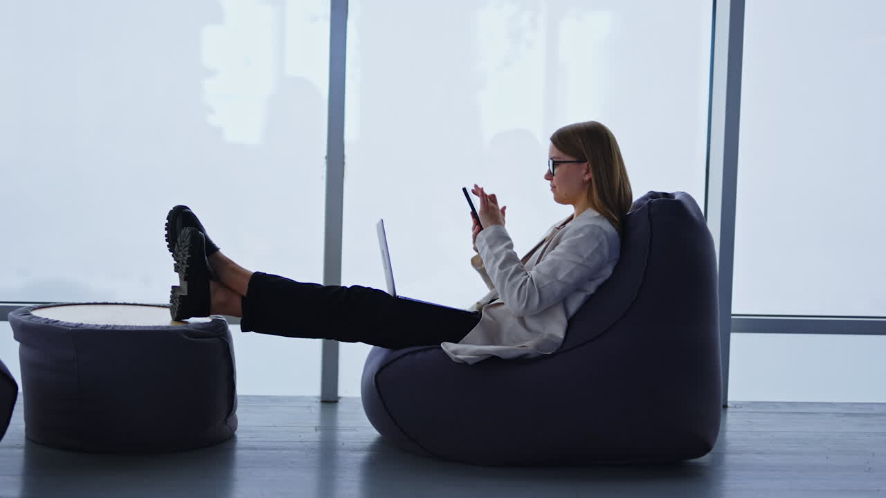 Lady in jacket and jeans distracted from work on her laptop. Young woman scrolling something on her phone. Panoramic window backdrop.