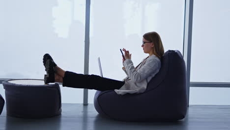lady in jacket and jeans distracted from work on her laptop. young woman scrolling something on her phone. panoramic window backdrop.