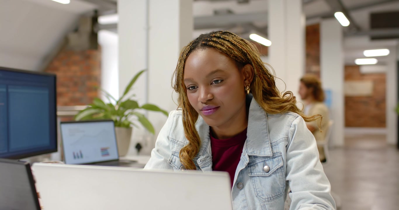 Female worker in denim jacket glancing at laptop leaning in studying at office as colleague passing