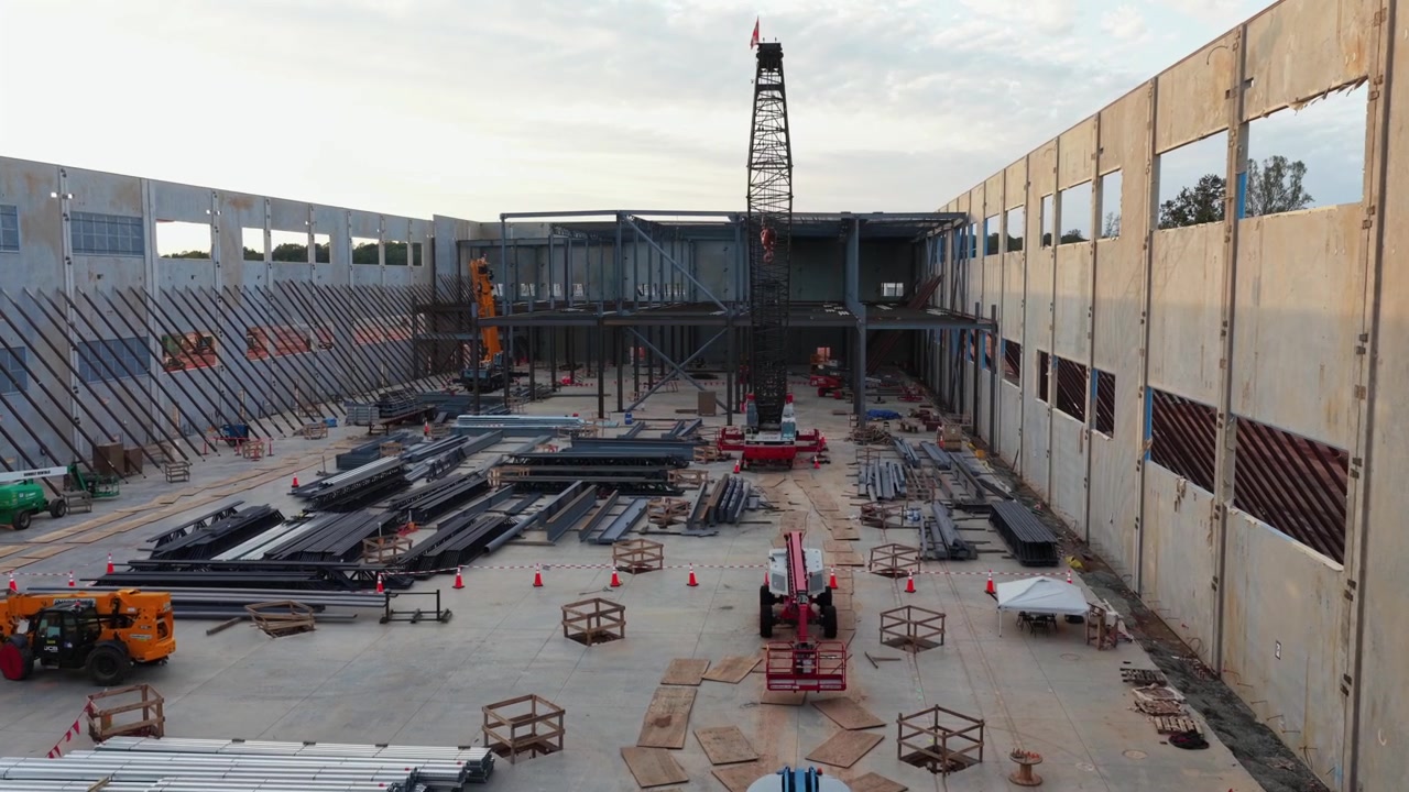 Wall opening in the tilt up panels of a data hall in Louisa County, Virginia, framing a crawler crane, stacked steel beams and boom lifts at work on the poured floor slab. Aerial approaching shot