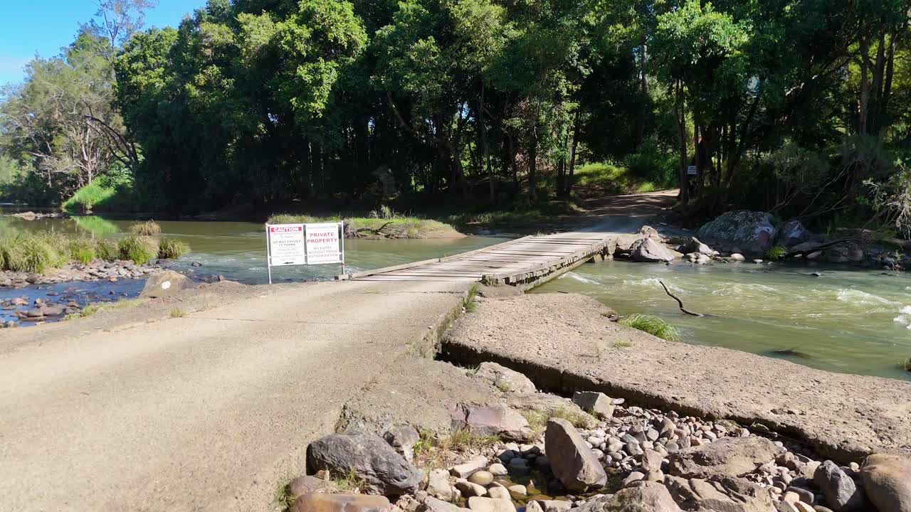 A dirt road crosses a creek with a private property sign. Surrounded by lush greenery under bright daylight