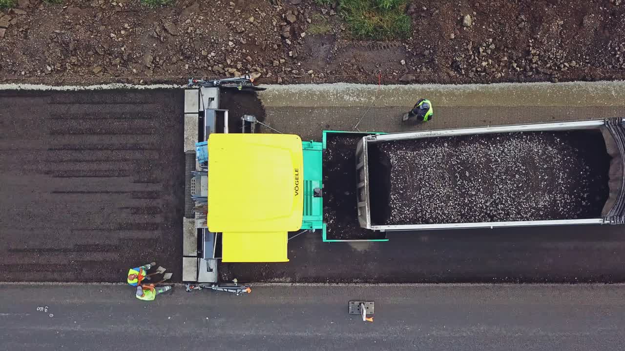 Black bitumen pours from the trailer of truck into the paver on the road on the background of repairmen in helmets and with shovels. Aerial view.