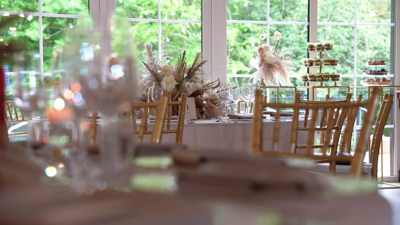 Meja resepsi pernikahan yang ditata dengan indah dihiasi dengan centerpiece bunga dan kursi emas. Cahaya sekitar yang lembut menciptakan suasana hangat dan mengundang dalam suasana elegan ini