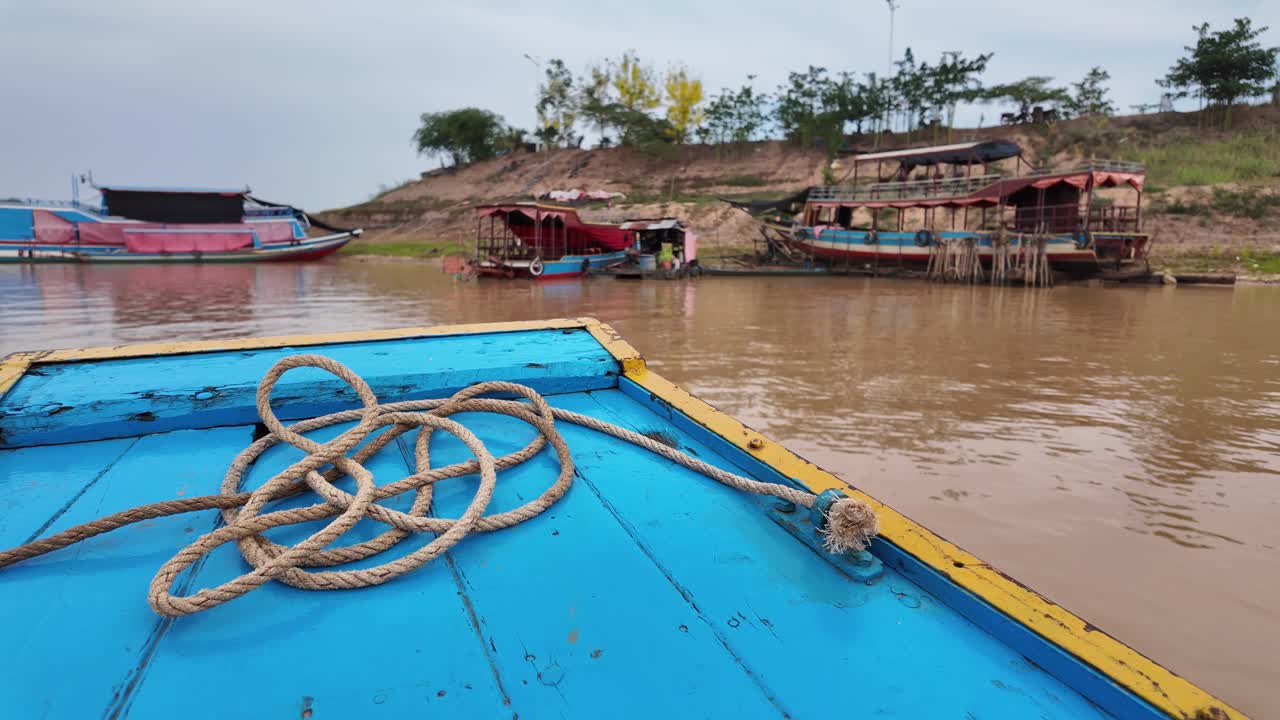 View from a blue boat on the muddy Cambodian river, with coiled rope in foreground and colourful houseboats docked on the sandy riverbank under an overcast sky.