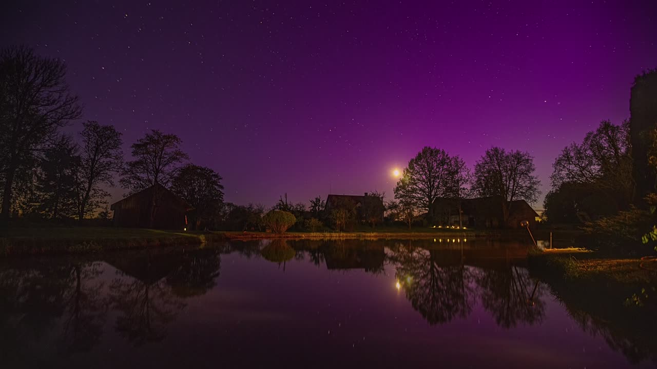 la luna se pone sobre una cabaña de campo junto a un estanque con estrellas relucientes y aurora boreal - lapso de tiempo nocturno