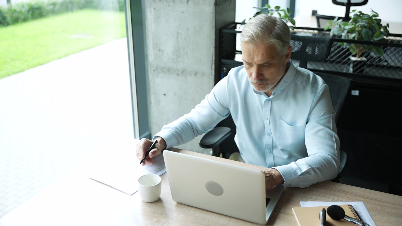 Mature Businessman Working on Laptop and Writing Notes in Modern Office