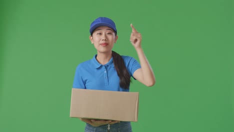 푸른 유니폼을 입은 아시아 여성 택배업자가 미소 짓고 스튜디오에서 녹색 스크린 배경에 상자를 배달하는 동안 카메라와 이야기합니다.