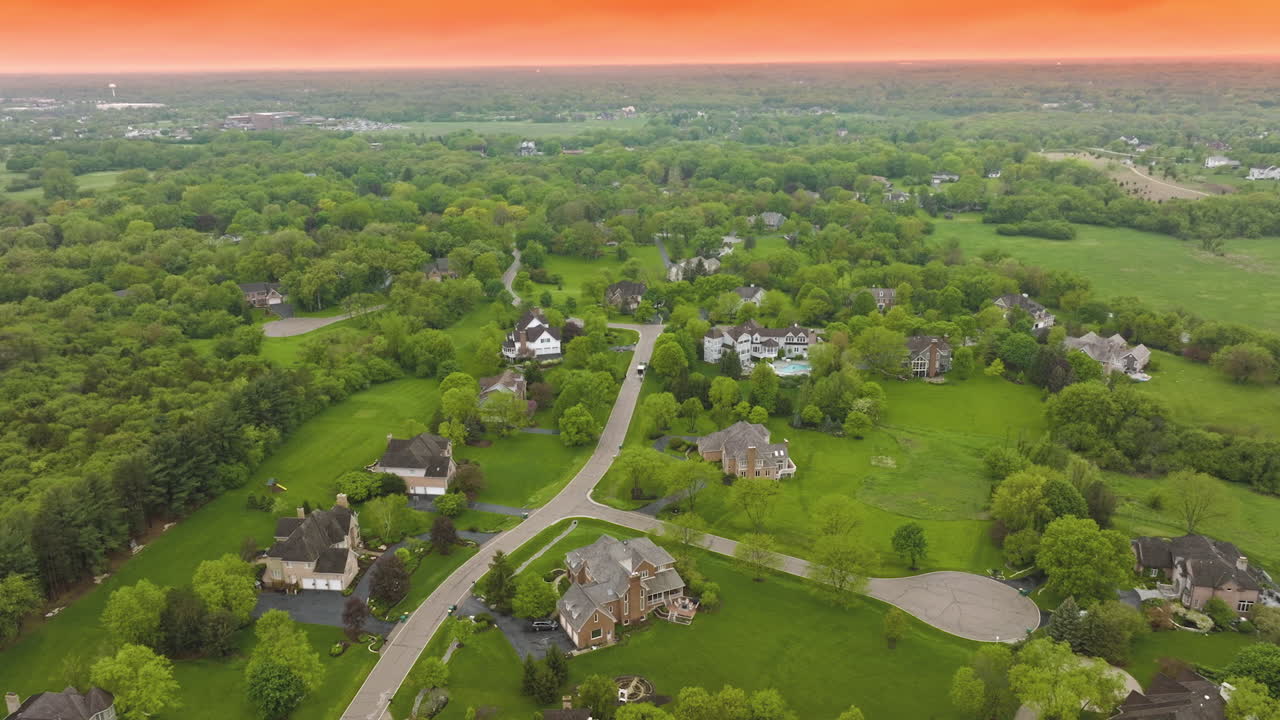 Well- kept suburban area in USA with beautiful private houses and neat roads. Rural neighborhood surrounded by green trees and grass lawns at the backdrop of orange sky.