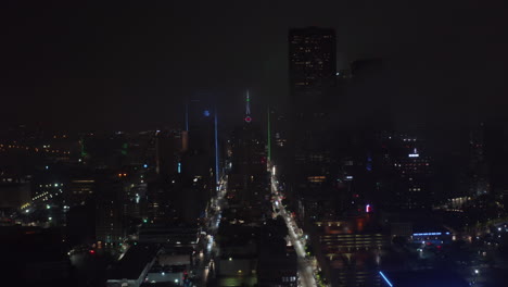 Drone-flying-over-illuminated-straight-street-between-tall-buildings-around-Mercantile-National-Bank-Building-with-tower-and-big-neon-clock.-Night-aerial-view-of-downtown.-Dallas,-Texas,-US