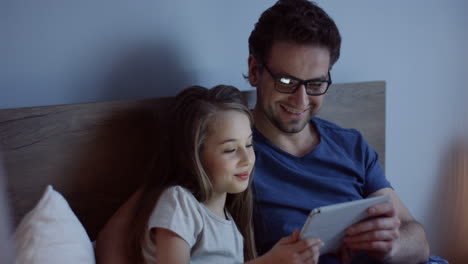 handsome caucasian man in glasses with his little pretty blond daughter watching something on the tablet and smiling while they sitting on the bed at night