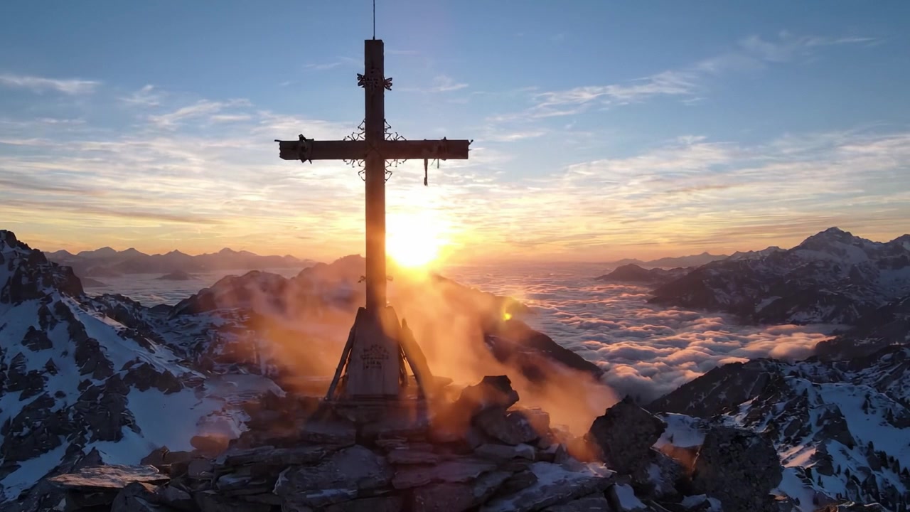 Fantastisk fjellsoloppgang med et kors: En fengslende utsikt fra toppen over et skyhav og majestetiske topper.
