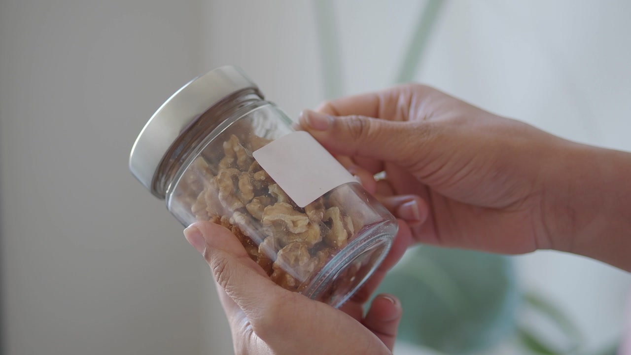 Hands Removing a Label from a Glass Jar of Walnuts