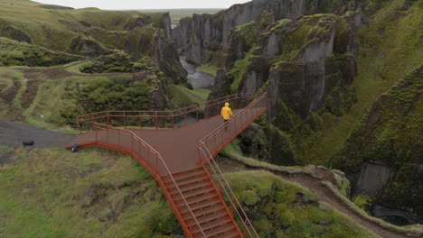 Antena:-Paso-Elevado-De-Una-Persona-Con-Chaqueta-Amarilla-Caminando-Sobre-La-Plataforma-Hacia-El-Cañón-Fjadrargljufur-En-Islandia