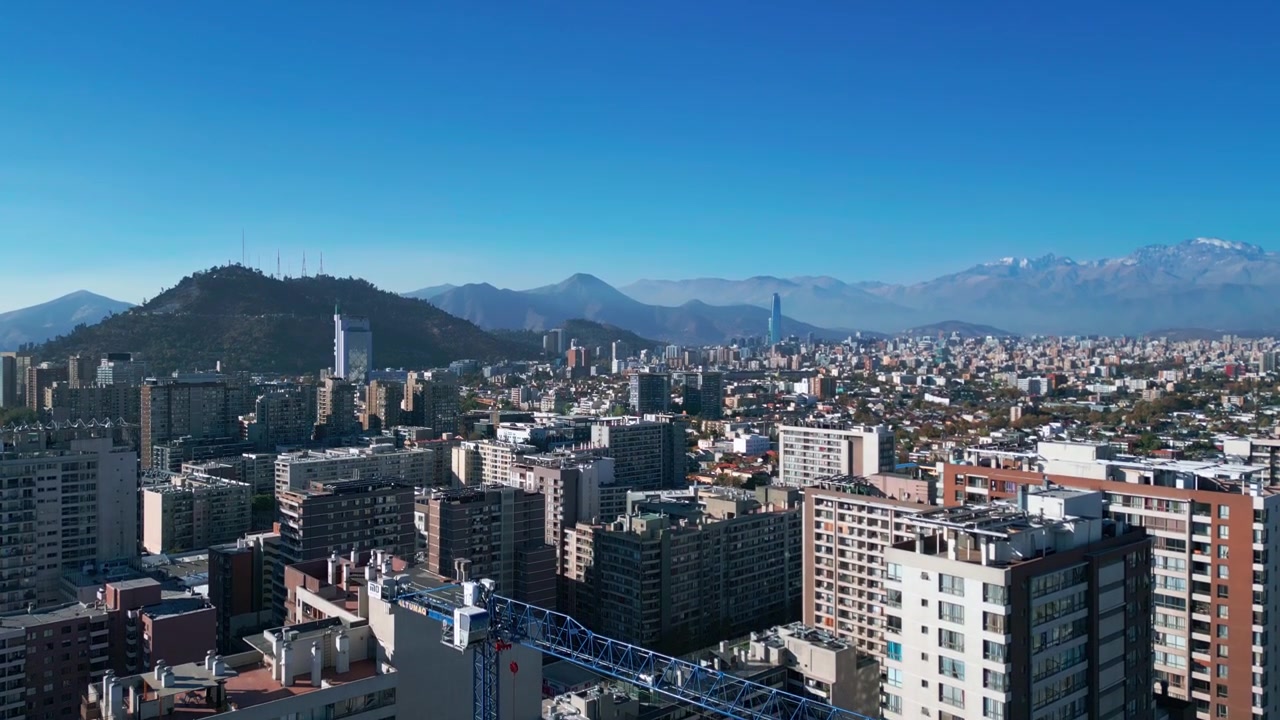Inquadratura ascendente su un denso quartiere residenziale urbano che rivela lo skyline della città con la torre Costanera Center, la collina di San Cristobal e le Ande innevate. Santiago, Cile.