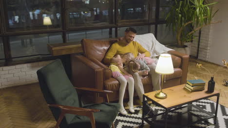 happy caucasian father sitting on sofa with his two sleepy little girls who leaning their heads on his lap