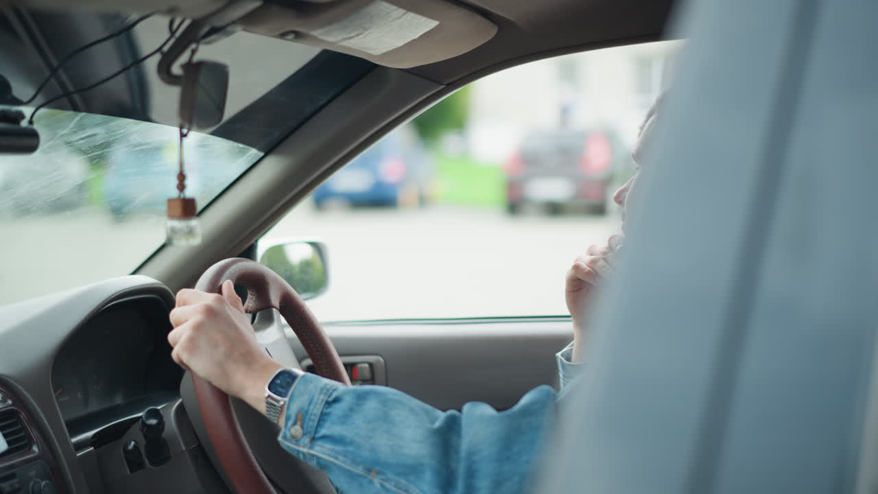 Varsle hvit sjåfør som skanner parkeringsplassen i denimjakke, med hånden på rattet mens han sjekker speil og blindsone, forbereder seg på å rygge eller flette inn i trafikken, bybakgrunn med parkerte biler, oppmerksom kroppsholdning