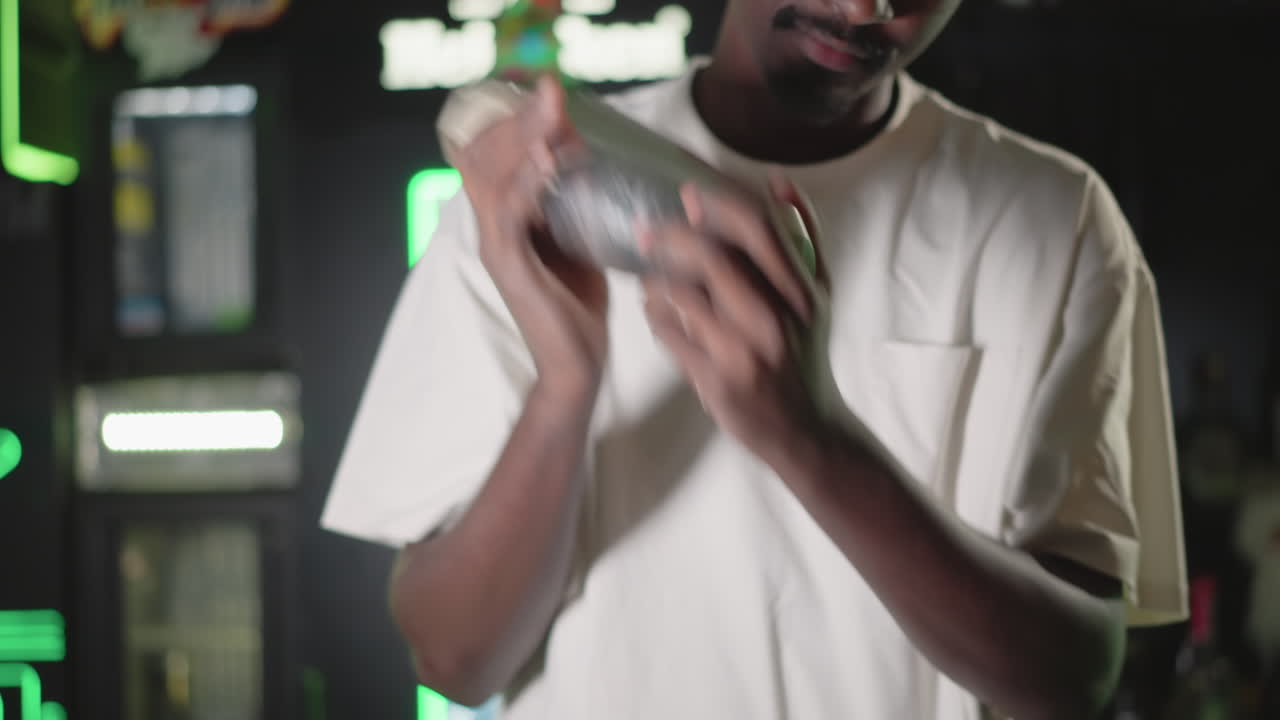 Bartender holding cocktail shaker with focus on hands, preparing drink in bar. Behind, illuminated liquor shelves and Heineken neon sign glow. Professional cocktail mixing in action