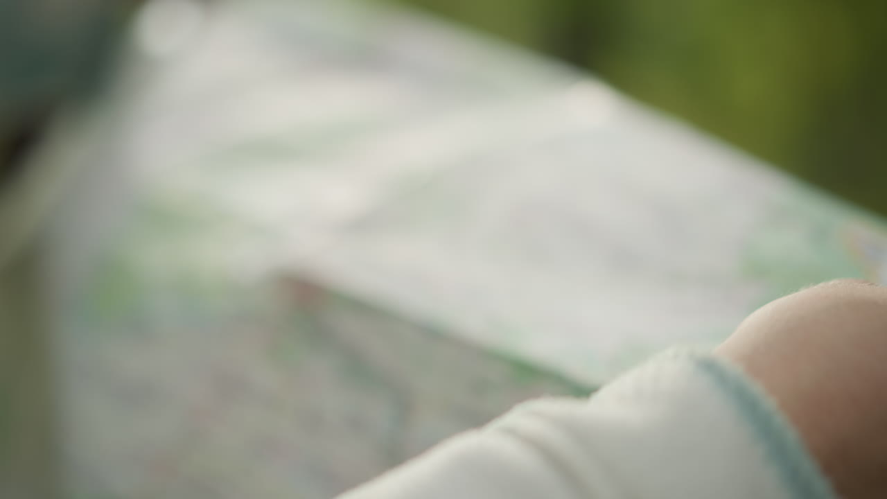 Nærbillede af en hvid vandrer, der folder et papirkort ud og følger ruter på et grønt topografisk print med hænderne, blød bokeh-skovbaggrund, morgenlys på krøllet papir, omhyggelig planlægning af solotur