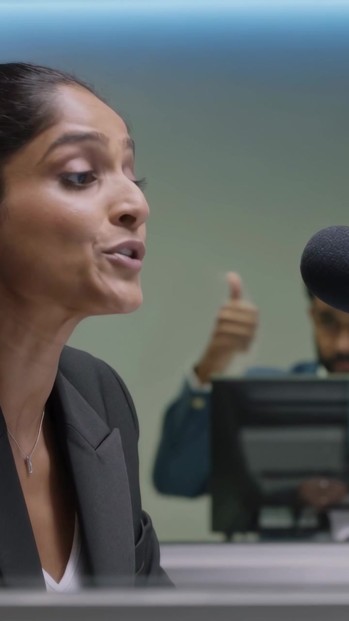 Vídeo vertical: Mulher de blazer inclinada gravando no microfone em mesa atrás do vidro, com produtor gesticulando.
